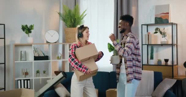 Хорошо выглядящая счастливая улыбающаяся молодая многонациональная пара, веселящаяся вместе в своей новой квартире с танцами и дающая друг другу пять — стоковое видео