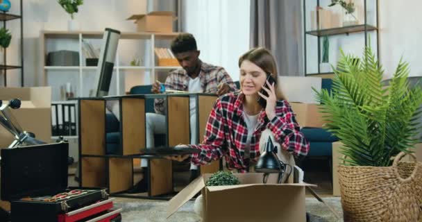 Likable confident young black-skinned guy measuring sizes of rack with tape measure when his cheerful pretty girlfriend unpacking cartons and talking on mobile,relocation concept — Stock Video