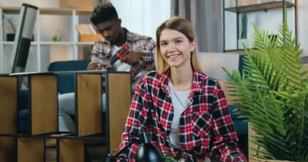 Close up of beautiful happy joyful blond girl which looking into camera with sincerely smile on the background of her black-skinned male friend which assembling rack with electric screwdriver — 비디오