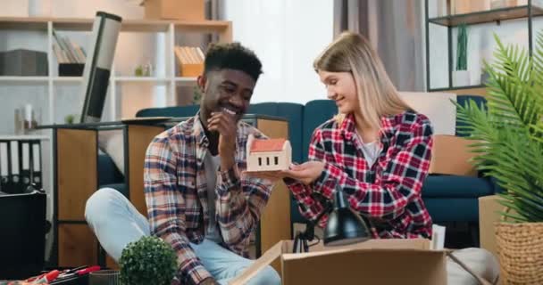 Likable felice sorridente spensierato giovane coppia di razza mista seduto sul pavimento nella loro nuova casa e disimballaggio cartone con elementi di arredamento — Video Stock