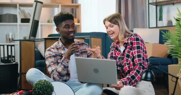 Vacker leende kreativ ung flicka dela med sig av sina idéer om att remodeling nytt rum tillsammans med hennes vackra positiva svarthyade pojkvän med hjälp av laptop — Stockvideo