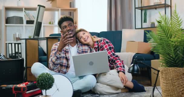 Mooi gelukkig glimlachend jong meisje zetten haar hoofd op zwart-gevilde vriendjes schouder en ze samen praten over het ontwerp interieur van hun nieuwe appartement met behulp van regeling op laptop — Stockvideo