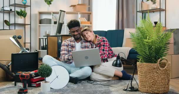 Probable heureux joyeux jeune couple multiracial assis sur le sol dans leur nouvel appartement entre différentes choses lors de la relocalisation et la conception de planification à l'aide d'un programme informatique spécial — Video