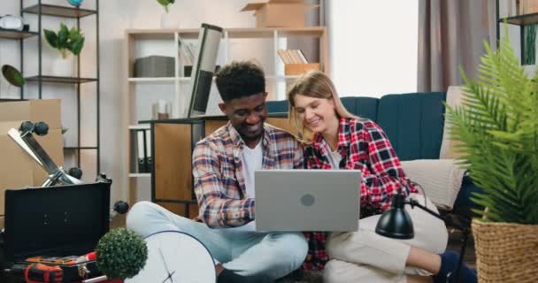 Adorable feliz joven y sonriente pareja de raza mixta moderna sentada entre cajas desempacadas en un piso nuevo y disfrutando de ver en la computadora, de cerca — Vídeos de Stock