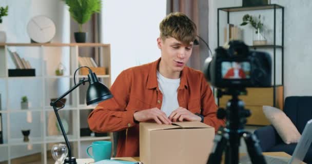 Концепция блога, где привлекательный современный молодой человек сидит перед камерой в домашней студии и записывает процесс распаковки для своего интернет-канала и показывает свои новые наушники для — стоковое видео
