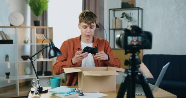 Guapo sonriente confiado joven moderno sentado frente a la cámara en la habitación acogedora y el proceso de desembalaje de grabación para sus seguidores y les dice acerca de joystick comprado — Vídeos de Stock