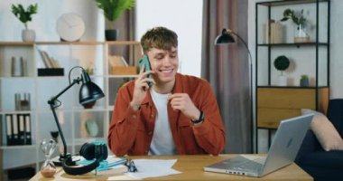 Çekici, güler yüzlü, koyu turuncu tişörtlü genç adamın portresi. Evde masada oturuyor ve arkadaşıyla cep telefonunda konuşuyor, ağır çekimde.