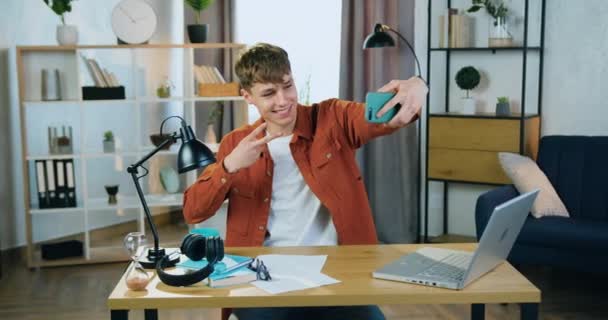 Probable sourire satisfait jeune homme insouciant faire selfie sur smartphone avec les doigts v-signe assis à la table dans une chambre confortable à la maison — Video