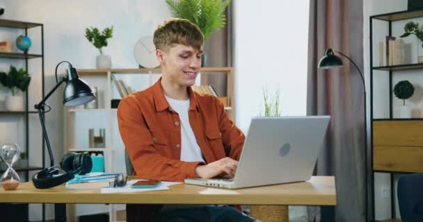 Atractivo chico joven trabajador positivo con camisa elegante sentado en su lugar de trabajo en la oficina en el hogar y trabajando en el ordenador portátil simultáneamente escuchar música de la lista de reproducción del teléfono — Vídeos de Stock