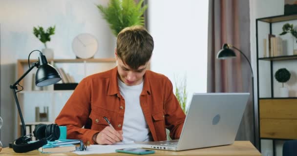 Handsome positive confident male student sitting in front of computer at home and listening online discussion from university teacher and making notes into papers — Stock Video