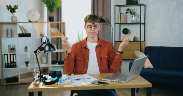 Concept de détente où beau jeune homme calme dans des lunettes assis à son lieu de travail dans son bureau à la maison dans la pose zen et méditer les yeux fermés en pause entre le travail — Video