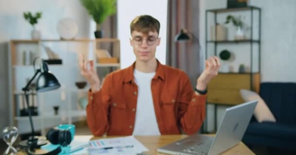 Caméra tournage sur beau jeune homme blond satisfait et détendu dans des lunettes qui gardent les mains mudra tout en méditant sur son lieu de travail dans un bureau à domicile confortable — Video