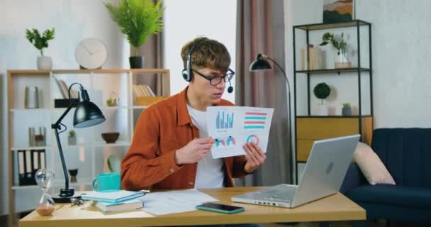 Vonzó, magabiztos, fiatal srác fülhallgatóban és szemüvegben, aki online videoértekezletet tart kollégáival, és vázlatos diagramokkal magyarázza a pénzügyi jelentés eredményeit. — Stock videók