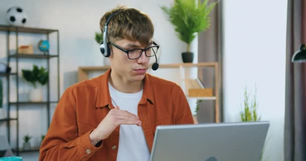 Provável confiante experiente jovem trabalhador do sexo masculino em fones de ouvido sentado na frente do laptop em casa durante o briefing on-line com colegas de trabalho e discutir detalhes do projeto conjunto — Vídeo de Stock