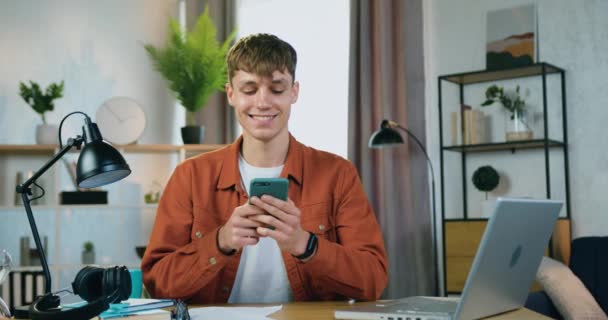 Atractivo feliz sonriente joven de moda sentado en la mesa en casa y utiliza su móvil para escribir mensaje a sus compañeros, concepto de ocio — Vídeos de Stock