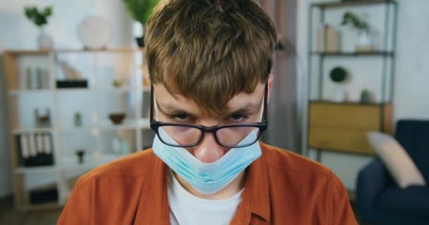 Zblízka mužský portrét v obličeji lékařská maska, která se dívá do kamery na pozadí útulné místnosti doma, ochrana koronavirového konceptu — Stock video