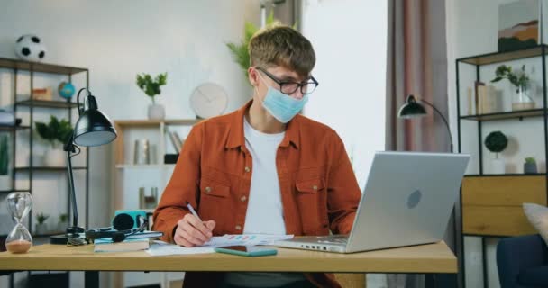 自宅のラップトップで働く顔の医療用マスクで魅力的な自信を持って勤勉な若い男性フリーランスや労働者、 covid-19 — ストック動画