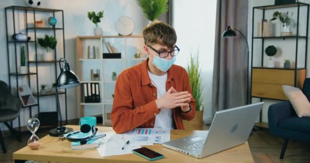 Retrato de un joven guapo y confiado en máscara médica que trabaja desde casa y habla con colegas o clientes durante la videoconferencia en el portátil, concepto coronavirus — Vídeos de Stock