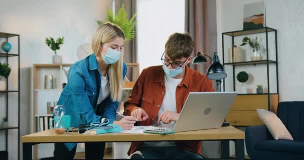 Atraktivní sebevědomý zkušený blonďatý pár v ochranných maskách spolupracující při startu používat informace o laptopu a dokumentech, koncepce práce s epidemickým koronavirem — Stock video
