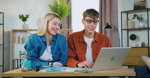 Pareja rubia joven satisfecha sonriente agradable que se sienta en la mesa en el país y utiliza el ordenador portátil para comprobar resultados de su trabajo o para encontrar la información necesaria — Vídeos de Stock