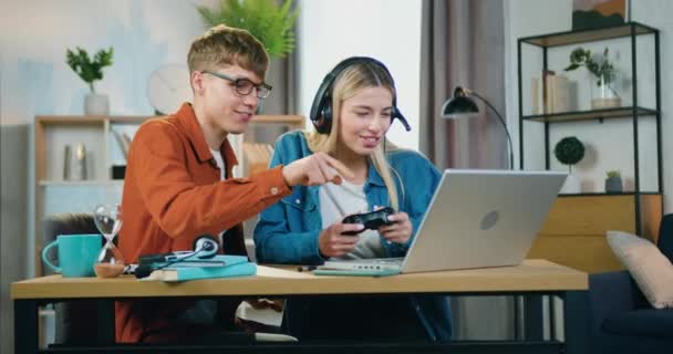 Atractivo sonriente despreocupado joven animando a su linda novia rubia alegre que se divierten mientras juega videojuegos en el ordenador portátil en casa, concepto de ocio — Vídeos de Stock