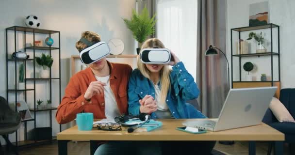 Primer plano de buena apariencia feliz pareja joven emocionada que se sienta a la mesa en casa y se divierte utilizando gafas de realidad aumentada, concepto de tecnología moderna — Vídeo de stock