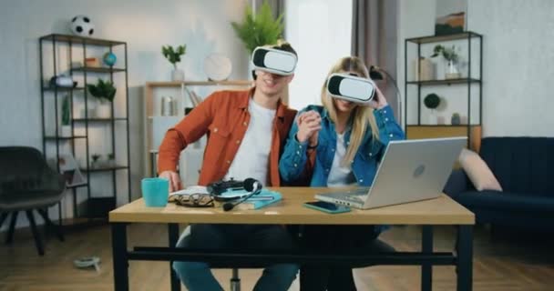 Camera shooting on good-looking smiling excited young couple in virtual reality headset which holding by hands and having fun during futuristic vision at home — Stock Video