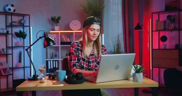 Schattig glimlachend tevreden jong blond meisje met pet eindigde te werken op laptop en zet haar handen achter het hoofd thuis in de avond, remote werk concept — Stockvideo