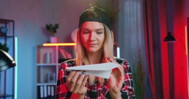 Zbliżenie przyjemny szczęśliwy szczęśliwy zrelaksowany blondynka młoda kobieta z kolczykiem w nosie, które trzymając papierowy samolot w rękach na tle przytulny pokój nocny — Wideo stockowe
