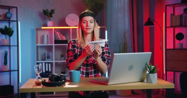 Привлекательная счастливая улыбающаяся современная молодая девушка с серьгой в носу, сидящая на рабочем месте дома вечером и запускающая бумажный самолет — стоковое видео
