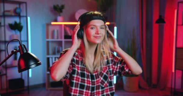 Φωτογραφική μηχανή γυρίσματα σε όμορφη χαμογελαστή ευτυχισμένη νεαρή ξανθιά κοπέλα στα ακουστικά που απολαμβάνουν μοντέρνα χαλαρωτικά τραγούδια σε όμορφα διακοσμημένο δωμάτιο με νυχτερινά φώτα — Αρχείο Βίντεο