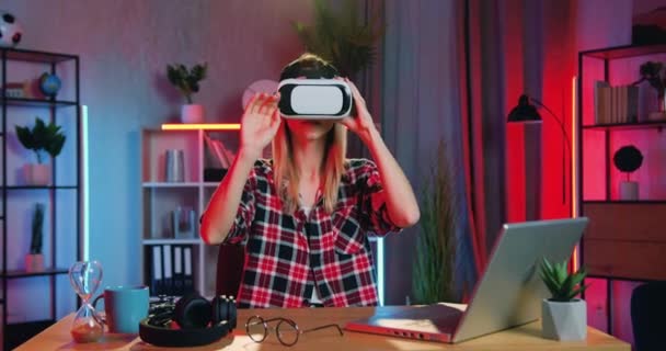 Concepto de tecnología moderna donde atractiva joven rubia inteligente y segura sentada en su lugar de trabajo en la oficina en el hogar por la noche y trabajando en auriculares de realidad virtual — Vídeo de stock