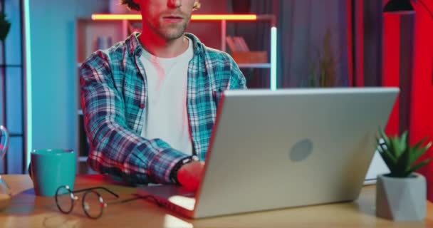 Красивый позитивный расслабленный молодой бородатый парень работает на ноутбуке на своем рабочем месте в домашнем офисе в вечернее время, концепция удаленной работы — стоковое видео
