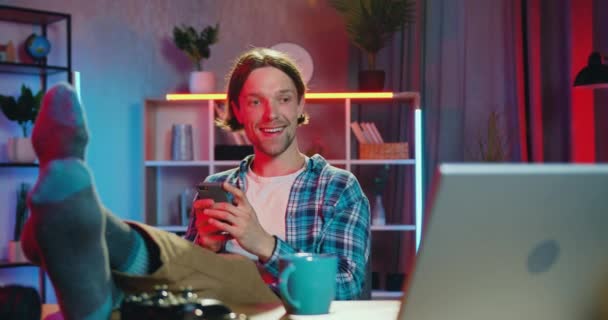 Likable joyful carefree young bearded man sitting in front of computer putting his legs on the desk at home in the evening during watching movie and using his mobile — Stock Video