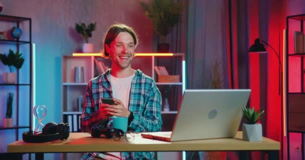 Портрет красивого улыбающегося беззаботного современного бородатого парня 25 лет, который сидит за столом дома вечером и смотрит комедию на ноутбуке одновременно печатая на смартфоне — стоковое видео