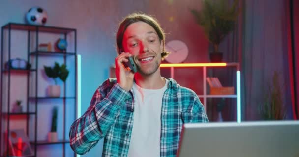 Beau sourire satisfait jeune homme barbu avec boucle d'oreille dans le nez tapant sur ordinateur portable lors d'une conversation mobile positive à la maison le soir — Video