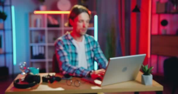 Camera shooting on handsome smiling skilled young bearded guy which sitting at his workplace in home office in front of laptop and looking into camera in the evening — Stock Video