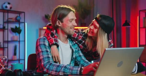 Krásná šťastná usměvavá zamilovaná dívka líbání na tvář její pozitivní přítel, zatímco oni spolu dělat on-line nakupování na počítači doma ve večerních hodinách — Stock video