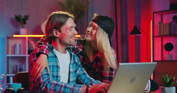 Lovely happy smiling blond girl hugging and kissing to the cheek her positive boyfriend when they together making purchases in online market on computer at home with night lighting — стоковое видео