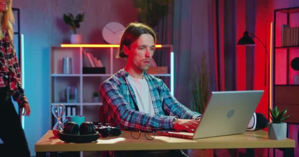 잘 생기고 믿음직 해 보이는 젊은 턱수염을 기른 젊은이가 저녁에 집에서 노트북으로 일하고 있습니다. 금발의 여자 친구가 그에게 와서 — 비디오