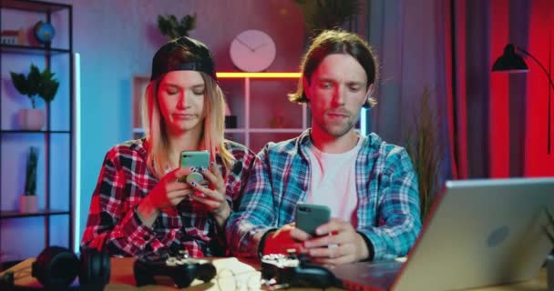 Atractiva pareja joven moderna reflexiva sentados juntos en la mesa en casa por la noche y todos ellos utilizan su teléfono inteligente — Vídeos de Stock