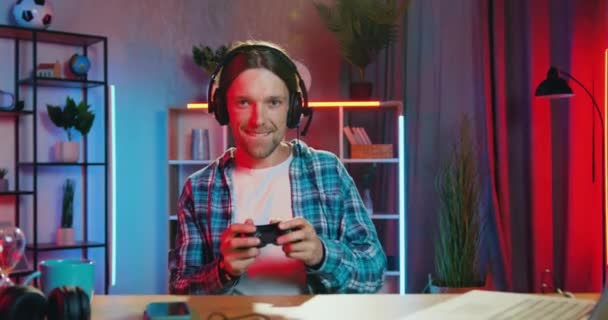 Mänskliga känslor koncept där stilig glada nöjda leende skäggig kille i hörlurar glädje från sin seger i videospel hemma på kvällen — Stockvideo