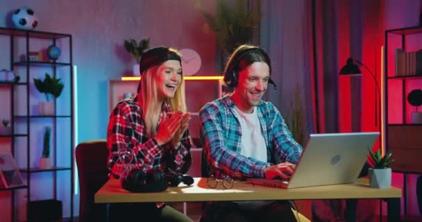 Jolie jeune blonde émotionnelle souriante soutenant son copain barbu excité actif qui joue à des jeux vidéo sur ordinateur portable à la maison le soir — Video