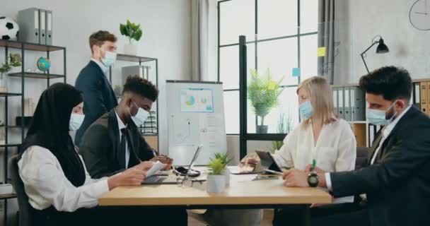 Werk in de tijd van covid-19 waar goed uitziende zelfverzekerde hoofdmanager nadert zijn bekwame multiraciale ondergeschikten in beschermende maskers en begroet ze met vuisten — Stockvideo