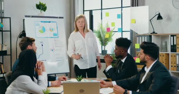 Atraktivní sebevědomá 45-letá blondýnka vedoucí drží prezentaci pro své pozorné smíšené rasy podřízených v moderní kancelářské místnosti — Stock video