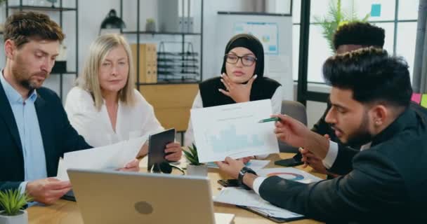 Concepto de trabajo de oficina donde atractivo hombre de negocios cualificado barbudo sentado en el lugar de trabajo junto con otros miembros del equipo de trabajo y explicando los resultados de su trabajo a los socios en línea — Vídeo de stock