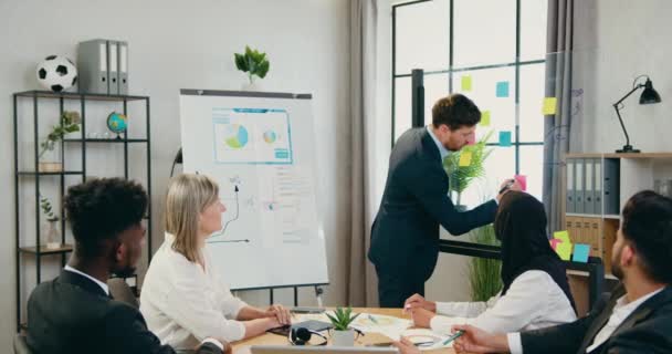 Conceito de trabalho de escritório onde boa aparência confiante equipe mista qualificada sentada no local de trabalho e assistindo como seu gerente principal barbudo bem sucedido escrevendo em notas pegajosas durante o briefing — Vídeo de Stock