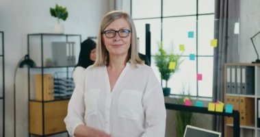 Güzel, güler yüzlü, kendine güvenen, gözlüklü sarışın iş kadını modern ofis odasında çapraz kollarıyla kameraya poz veriyor.