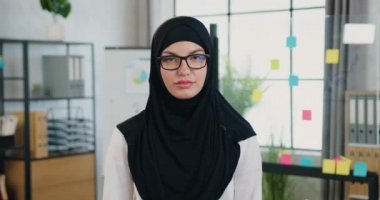 Büyüleyici gülümseyen, başarılı, tesettürlü Arap iş kadını modern ofis odasının arka planındaki kameraya bakıyor.