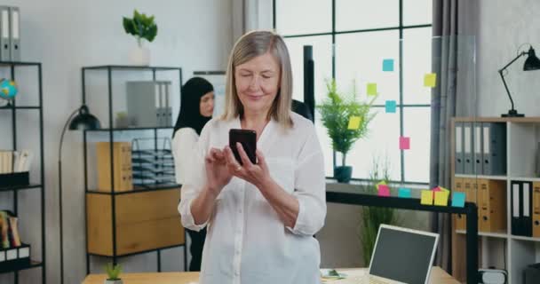 Vonzó magabiztos pozitív nyugodt 45 éves szőke üzletasszony áll a közepén a modern iroda és használja az okostelefon — Stock videók